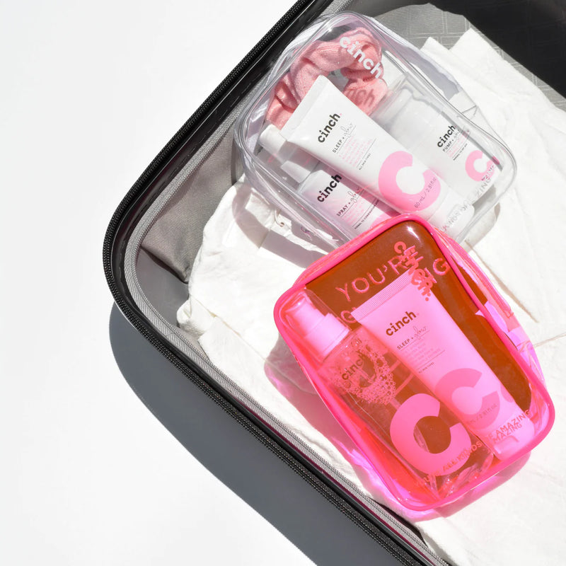 Cinch Skin Clear PVC Bag pictured in suitcase next to Neon Pink PVC bag. Both bags are filled with Cinch skincare products.